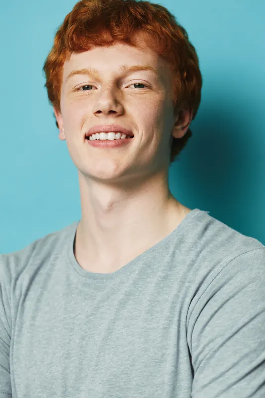 Teenager with red hair and smiling face wearing gray t shirt against blue background. Portrait of happy young man with casual style looking at camera in studio.