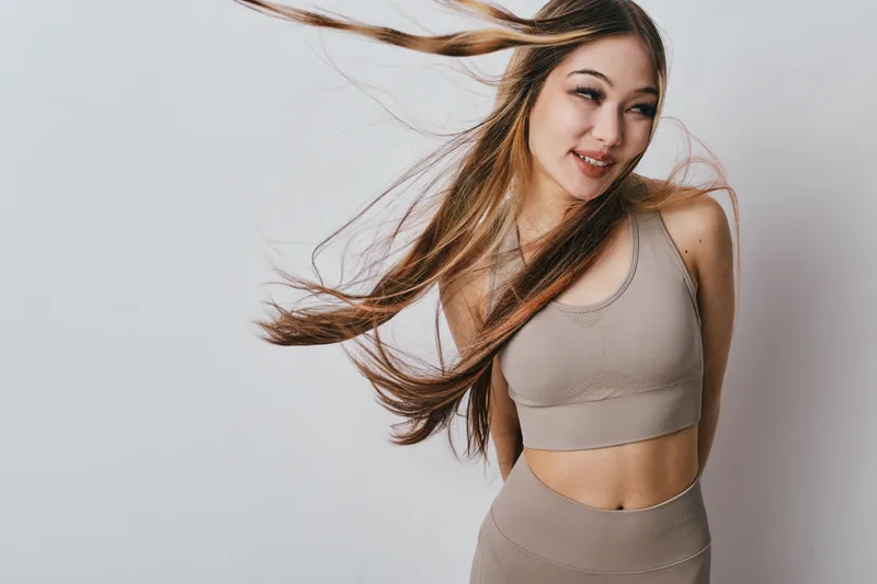 Young woman with long hair wears beige sportswear smiling with wind blowing hair posing against light background for fitness or lifestyle concept.