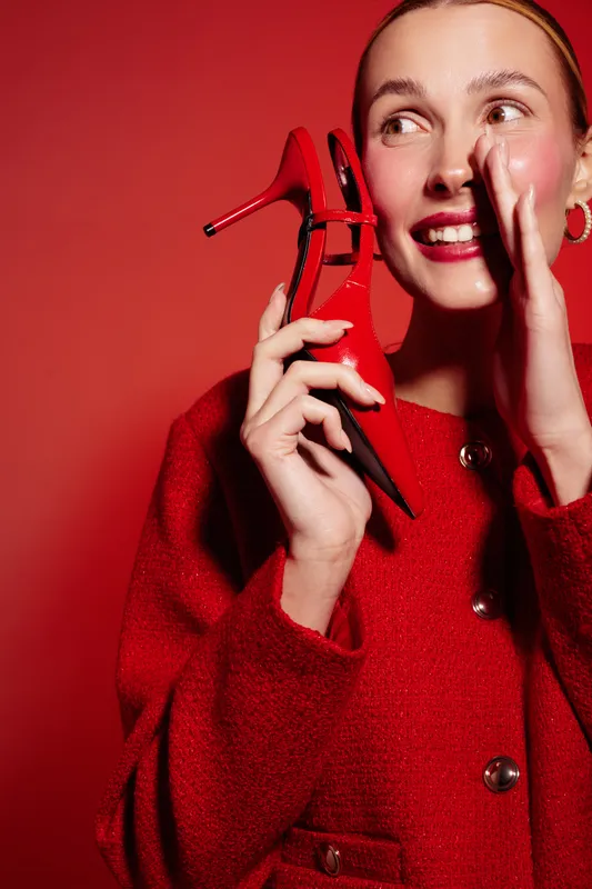 Smiling woman in red coat holds red high heel shoe near face, posing on red background. Concept of fashion, style, elegance, and vibrant color contrast in studio portrait photography.