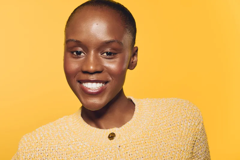 Smiling young black woman with dark skin wearing a yellow sweater stands against a bright yellow background. Concept of happiness, beauty, confidence, and positive emotions.