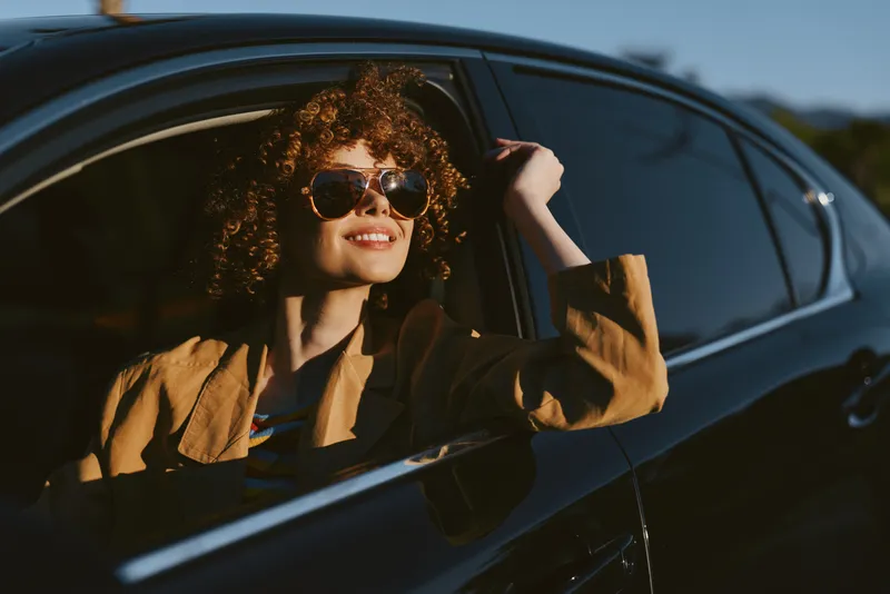 Cheerful woman in a car, wearing sunglasses, enjoying a sunlit moment, relaxed pose, warm retro film color, gentle breeze, candid outdoor lifestyle shot.