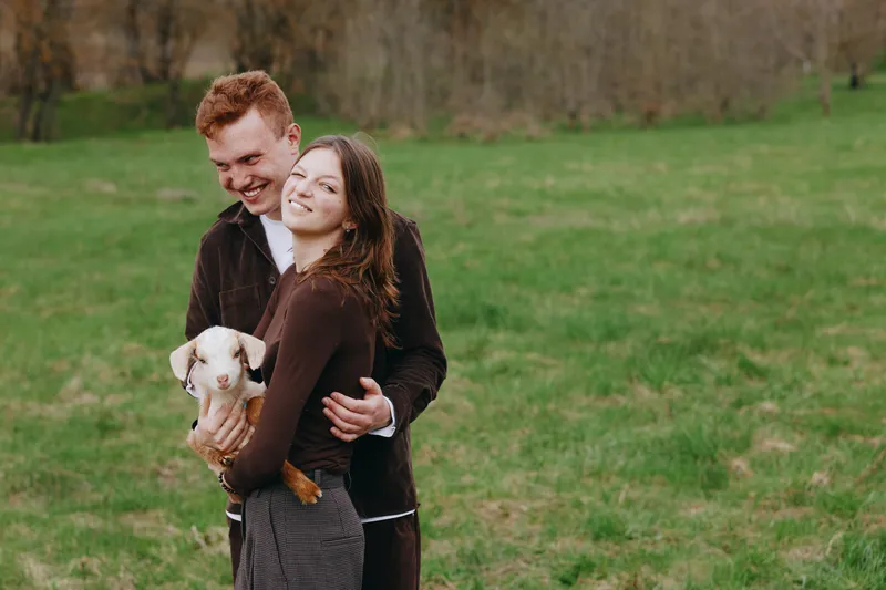 Joyful couple outdoors with a playful goat kid in a grassy field. Warm smiles, natural daylight, rustic vibe, and a wholesome lifestyle moment. Connection, care, and simple happiness in nature