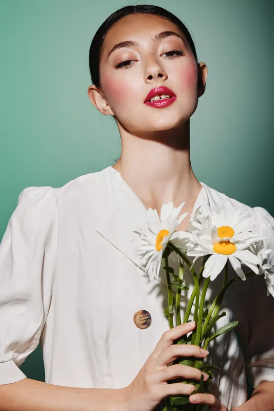 Young woman with bold makeup poses holding white daisies. Soft lighting enhances her flawless skin and creates an artistic vision of beauty and fashion creativity.