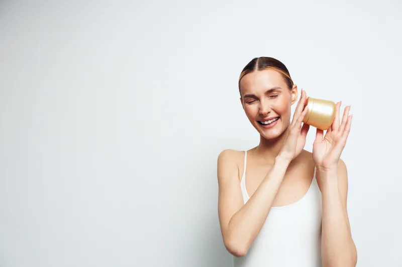 Young brunette woman in a white tank top smiles playfully while holding a roll of tape to her ear, posing with a light, neutral studio background in a candid, cheerful moment.