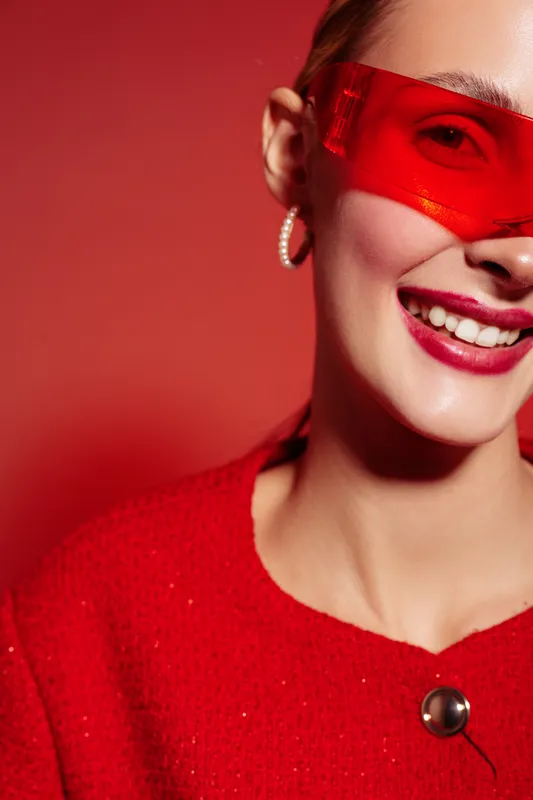 Smiling woman in red sweater and red translucent glasses poses on red background. Close-up portrait with bright makeup and hoop earrings. Concept of fashion, style, and confidence.