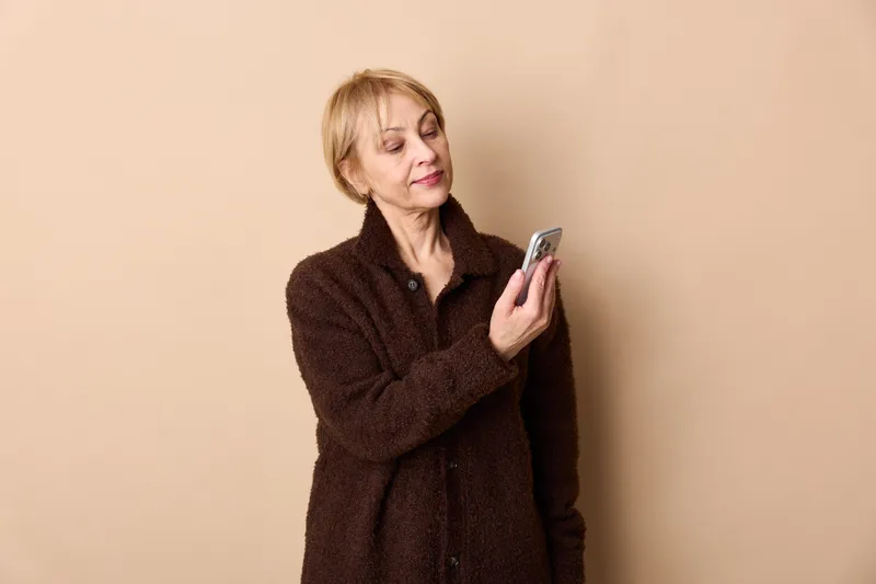 Elderly woman smartphone technology communication portrait indoors beige background, holding phone in hands, looking at screen with thoughtful expression, soft studio lighting, concept of digital