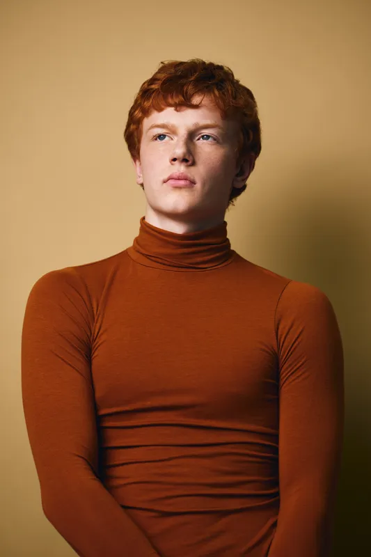 Young man with red hair and fair skin wearing a brown turtleneck sweater looking away with serious expression on neutral background. Portrait of confident male with natural light.