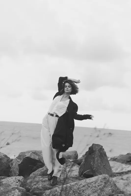 Monochrome fashion on a rocky shoreline, wind-swept pose with a flowing coat and dress, bold minimalist style against the sea backdrop.