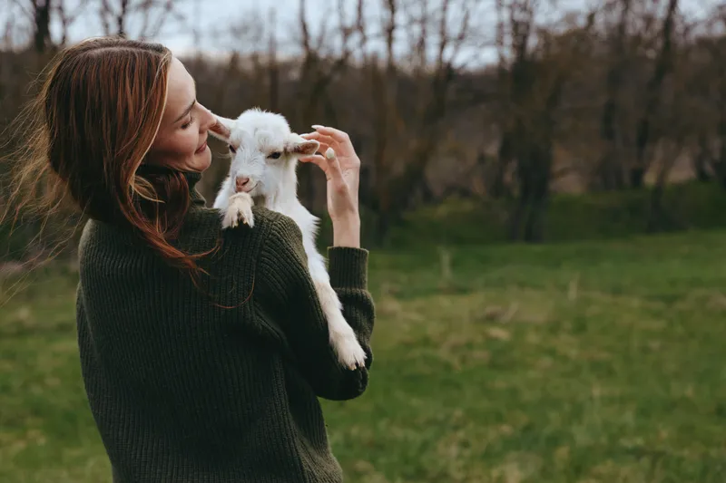 A woman outdoors holds a goat kid in a gentle hug, smiling softly in a grassy field. A candid moment of affection, calm bonding, and rural outdoor life.