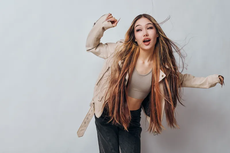 Young woman with long braided hair dancing and enjoying herself wearing casual beige jacket and black pants on gray studio background with dynamic pose and expression.