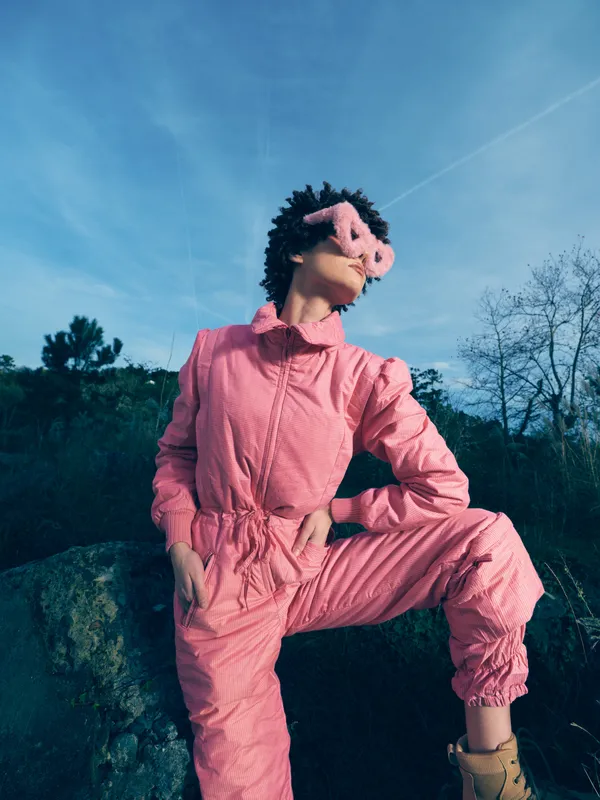 Outdoor portrait of a person in a pink jumpsuit wearing a pink masquerade mask, posing confidently on a rocky ledge under a clear sky. Fashionable street style with bold color and playful disguise.