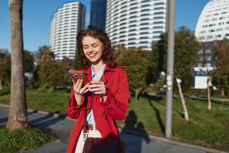 Smiling woman with curly hair wearing a red jacket looking at a smartphone outdoors. Urban park setting with modern buildings in the background wellness lifestyle
