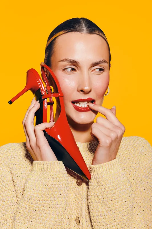 Young woman in beige sweater holds red high heel shoe near face, looking sideways with playful expression on bright yellow background. Concept of fashion, style, and beauty.