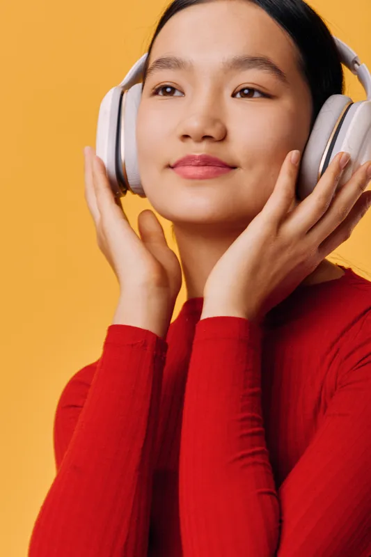Young woman in red sweater wearing wireless headphones enjoying music with closed eyes against orange background. Relaxation, technology, and sound experience with modern wireless device.