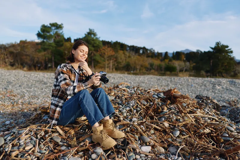 Photographer woman sitting on a rocky beach with a camera, wearing plaid shirt and boots, capturing a calm autumn shoreline with pebbles and distant trees under a clear sky.