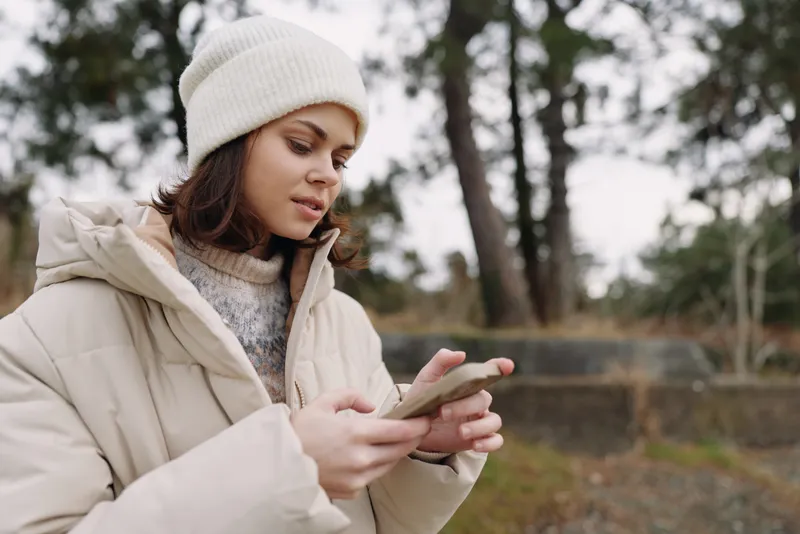 Woman in a beige jacket and white beanie outdoors, using a smartphone with a focused expression. Winter scene with blurred trees, calm mood, and chilly air, minimalist lifestyle shot.