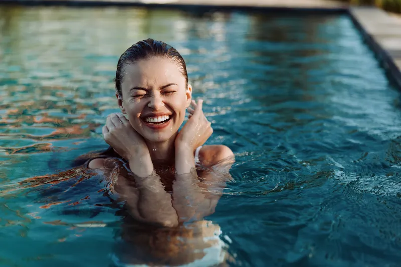 Joyful woman enjoys a refreshing dip in a bright pool, smiling with hands on cheeks, capturing a carefree summer moment of relaxation, water fun, and sunshine