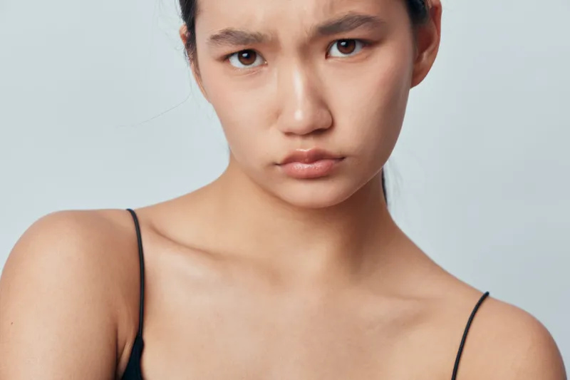 Portrait of young woman with serious expression in minimal clothing isolated on light background. Closeup face and shoulders with natural skin tone and intense look for emotion concept.