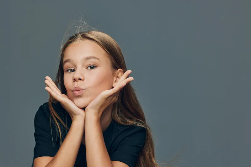 Girl with long hair in black shirt making playful face with hands under chin on grey background. Child posing and expressing joy with cute gesture indoors studio portrait.