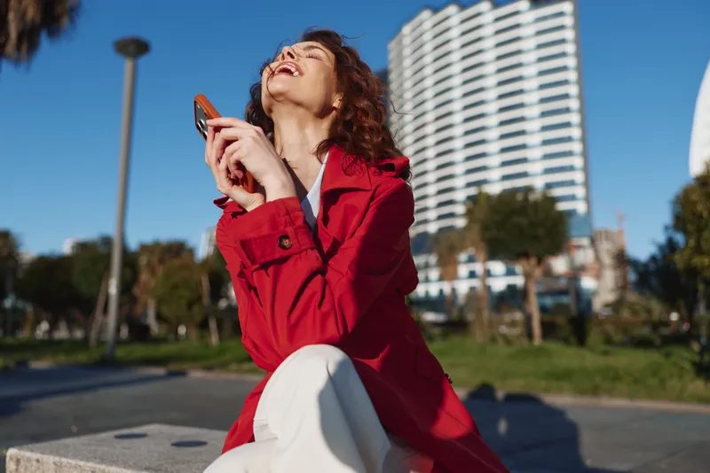 Young woman in a red coat laughs and looks up with a phone in hand, enjoying a sunny day in a bustling urban park beside modern high rise buildings.