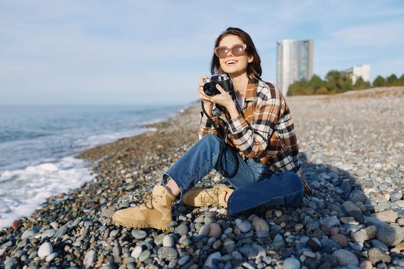 Woman photographer on a pebble beach with a camera, wearing a plaid shirt, denim jeans and boots, enjoying sunny coastal scenery and open sea view at the shore.