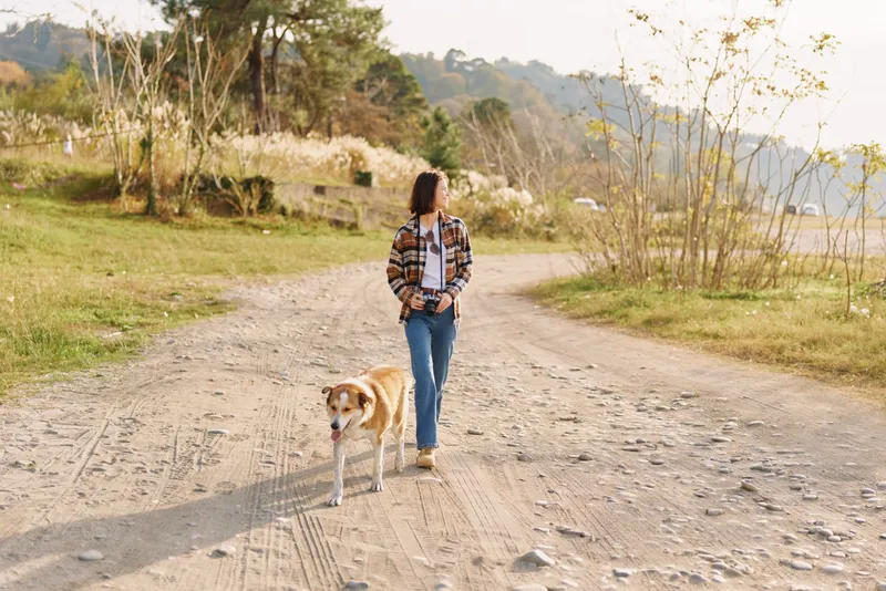 Woman walking a dog on a dirt road through a rural landscape, casual outfit, relaxed mood, golden hour light, trees and hills in the background, outdoor lifestyle moment.