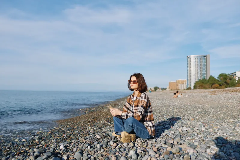 Beach scene with a woman sitting on pebbles by the ocean, wearing a plaid shirt and sunglasses, enjoying a quiet outdoor moment by the water.