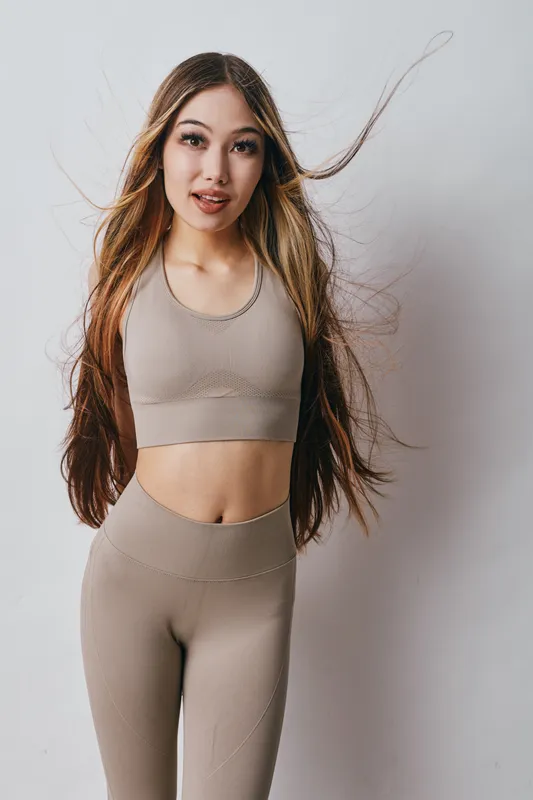 Young woman with long hair wearing beige sportswear standing against white background, fitness outfit, casual pose, healthy lifestyle concept, studio portrait.