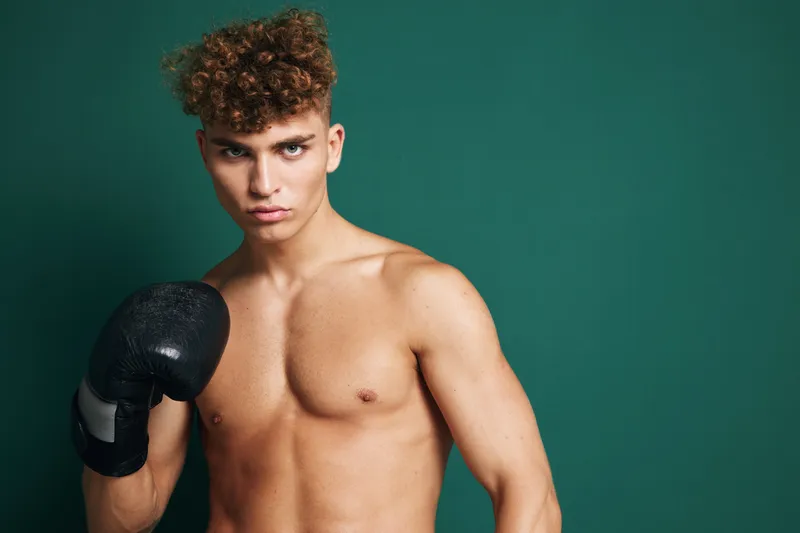 Young man with curly hair and boxing glove in training pose against dark green background expressing focus, strength and athleticism with bare skin above waist.