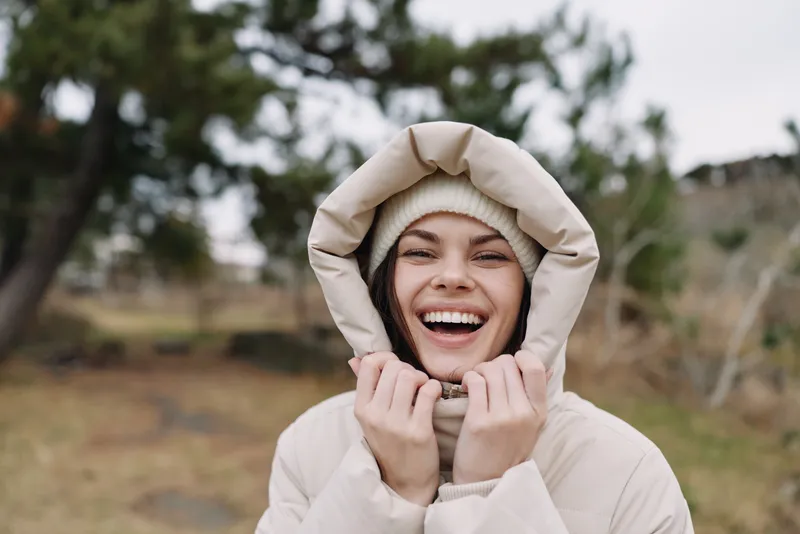 Woman in beige winter coat smiling outdoors with hood up, surrounded by nature, cozy and joyful portrait capturing bright mood in chilly weather