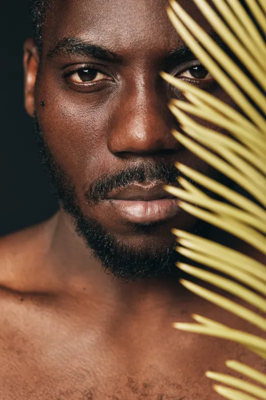 African man with dark skin and beard in creative conceptual visual style closeup portrait with palm leaf on black background. Artistic expression and natural beauty in studio lighting.