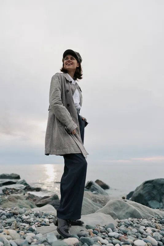 Young woman in a grey coat and hat stands on rocky beach near calm sea under cloudy sky. She looks happy and relaxed enjoying outdoor nature, casual fashion and peaceful mood.