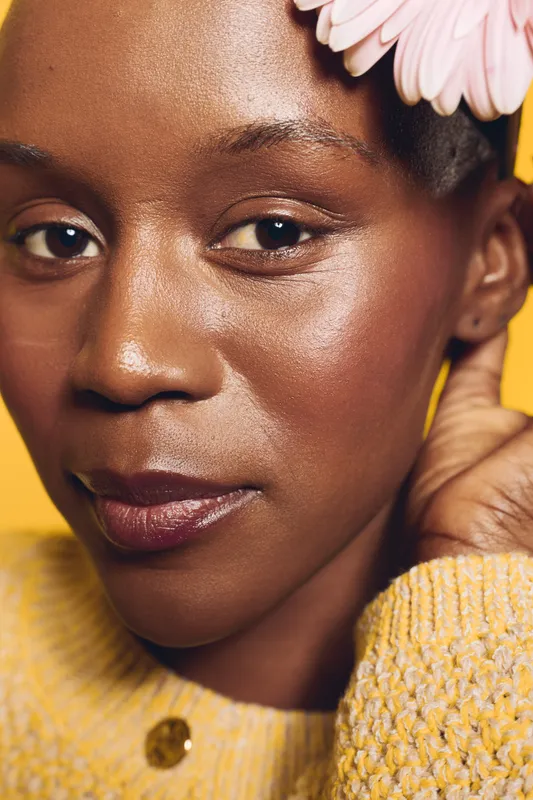 Close-up of an African American woman with dark skin wearing a yellow sweater and pink flower in hair. Natural beauty and skincare concept with warm lighting on yellow background.