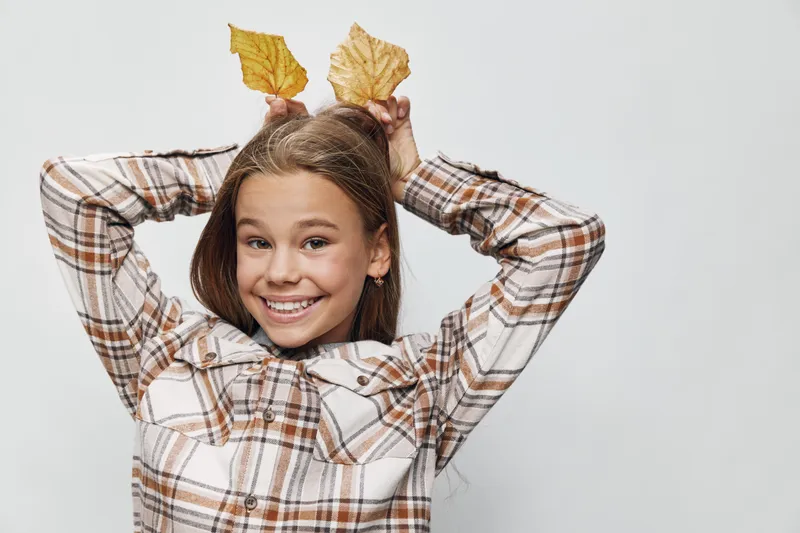 Girl smiling and holding autumn leaves above head with plaid shirt against white background. Child playing and posing for fall seasonal fun with brown hair and happy expression.