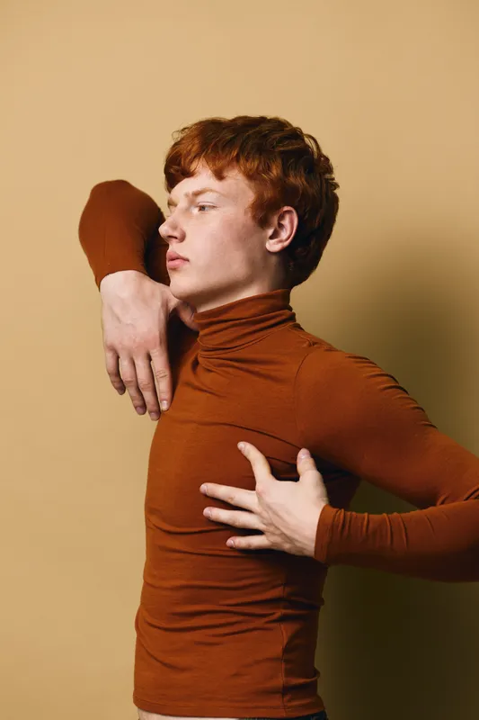Redhead man with fair skin and curly hair wearing brown turtleneck in minimal beige studio. Portrait with arm raised and side profile in artistic fashion pose and serious expression.