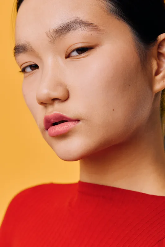 Portrait of young woman with smooth skin and red sweater looking at camera on yellow background. Closeup, natural makeup, serious expression, fashion and beauty concept in studio light.