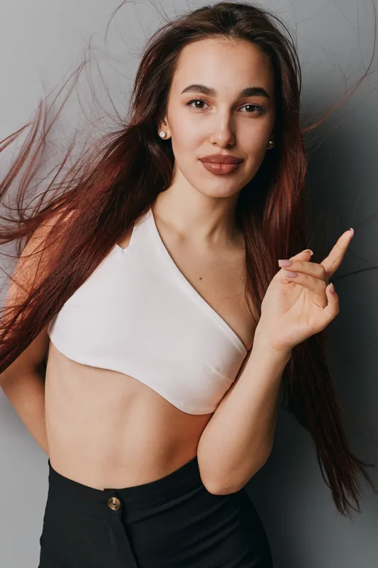 Young woman with long brown hair wearing white crop top and black pants posing with confident expression and finger pointing up against gray background in studio.