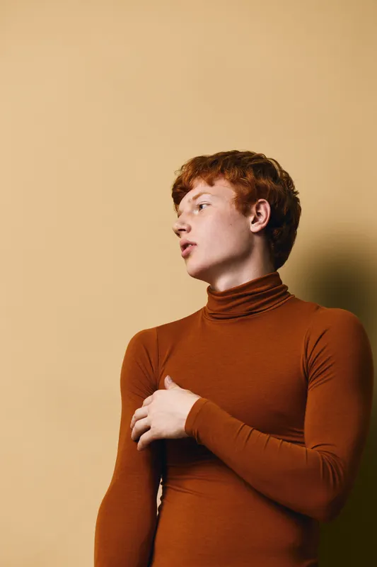 Young man with red hair wearing rust turtleneck sweater posing against beige background, looking sideways with contemplative expression in casual minimal studio portrait.