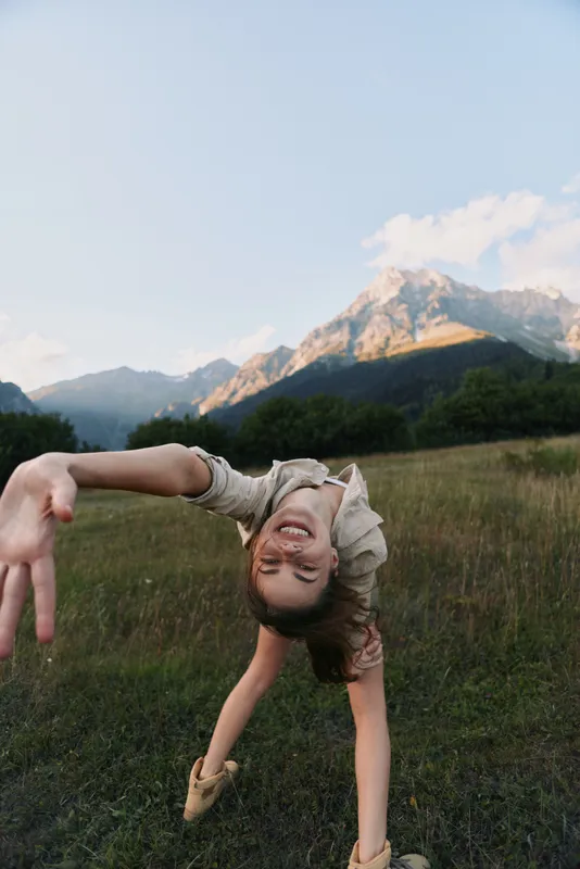 Woman outdoors in a meadow with mountains in the background performs a dynamic backbend, exuding playful energy, balance, and a joyful mood in a natural setting.
