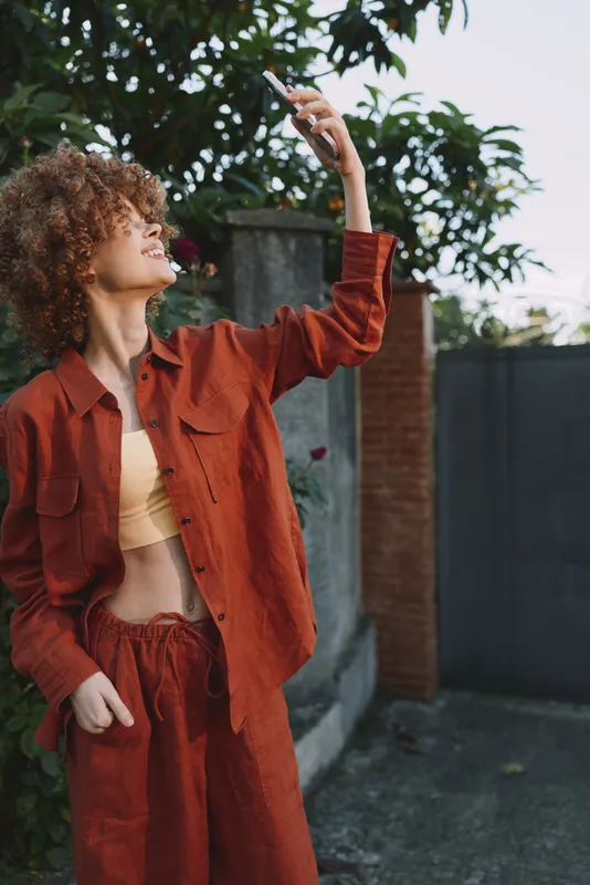 Cheerful woman outdoors wearing a rust red suit, cropped top, posing with one arm raised toward the sun, soft breeze, vibrant natural colors, relaxed lifestyle moment