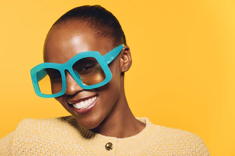Smiling dark skinned African woman wears oversized blue sunglasses and yellow sweater, posing against bright yellow background. Concept for fashion, summer, and joyful mood portrait.