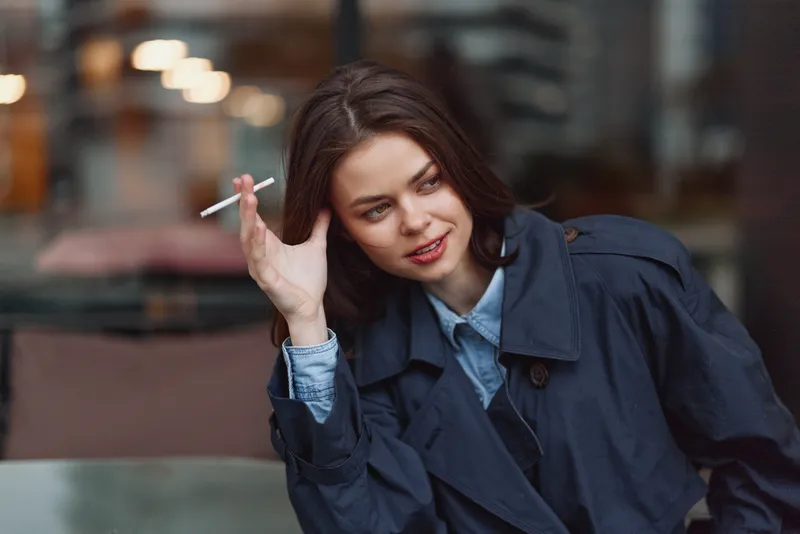 Casual young woman in a navy trench coat smokes a cigarette, leaning on a cafe table with a relaxed half smile as a blurred urban street frames the scene.