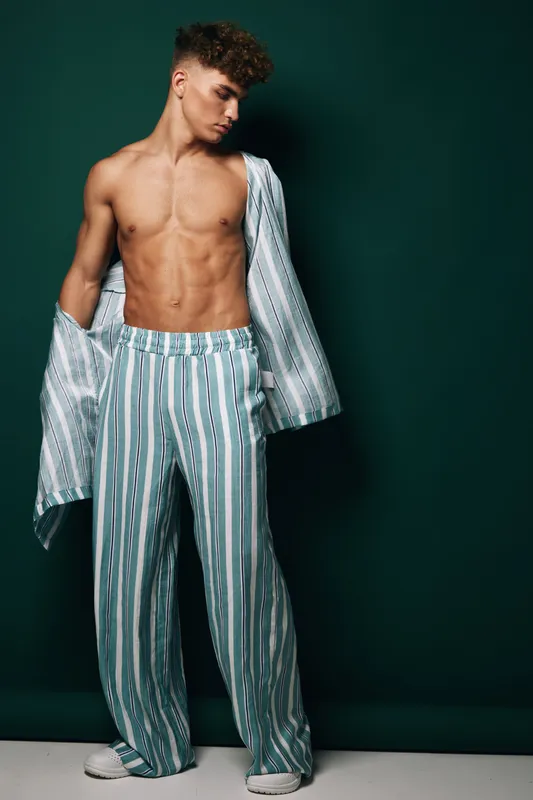 Model, young man, standing shirtless with an open striped shirt and matching wide trousers against dark green background, looking thoughtfully to the side exhibiting fit physique and fashion.