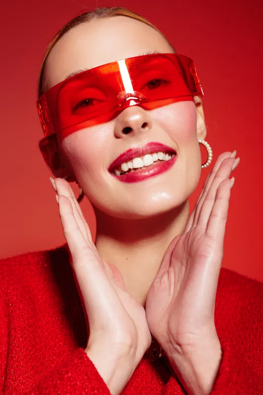 Smiling woman with red sunglasses and red lipstick poses with hands near face on red background. Fashion portrait with bright colors and confident expression. Concept of style and beauty.