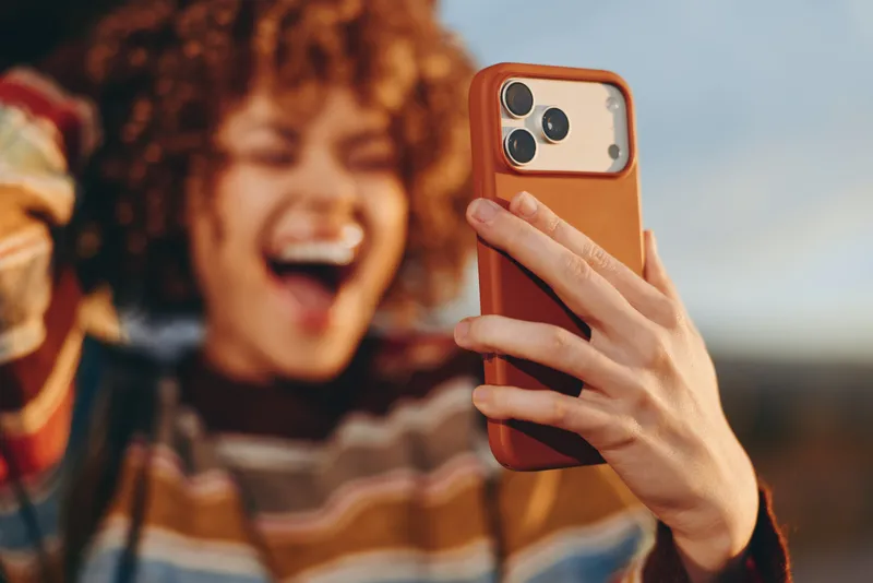 Woman lifestyle selfie with smartphone in hand and orange phone case outdoors at sunset. Smiling woman in background laughs with friends, casual mood, shallow depth.