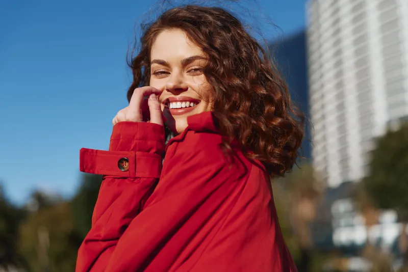 Young woman with curly hair smiles brightly wearing a red coat outdoors on a sunny day in an urban environment.