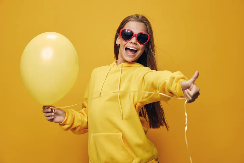 Young woman in yellow hoodie holding yellow balloon with happy smile and heart shaped sunglasses on bright background for celebration and joy mood.