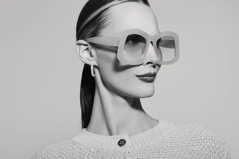 Portrait of a white woman with pale skin wearing large sunglasses and a textured sweater, looking sideways in a minimal studio setting.