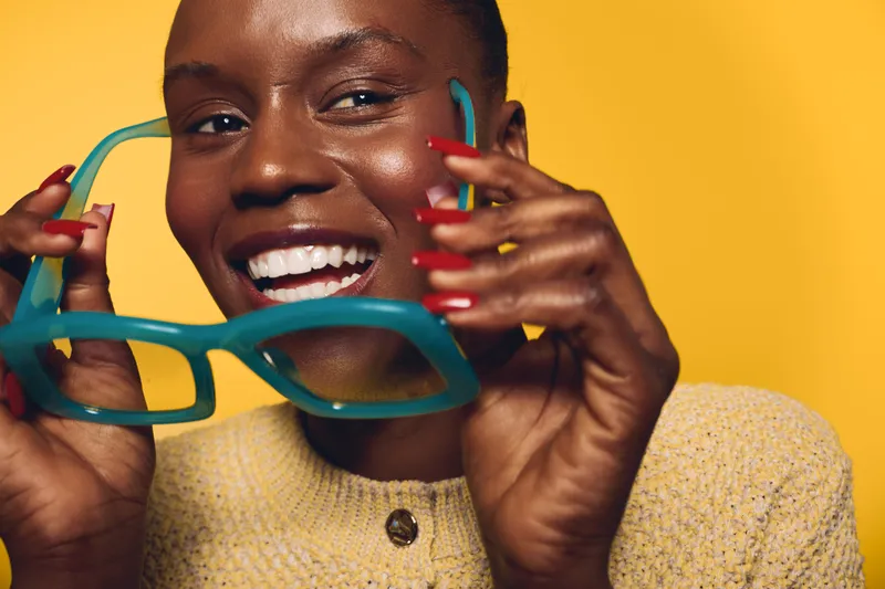 Happy dark skinned African woman with shiny skin holds turquoise glasses near face smiling brightly. Yellow background and red nails create vibrant and joyful atmosphere of beauty and fashion.