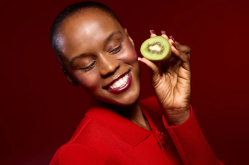 Smiling black African woman with dark skin holding fresh kiwi fruit near face on red background. Concept of healthy lifestyle, natural food, wellness, and vibrant mood in studio lighting.